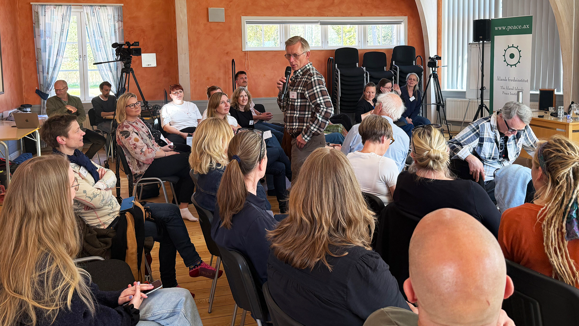 The picture is showing a room full of sitting people and one man speaking on the microphone in the middle.