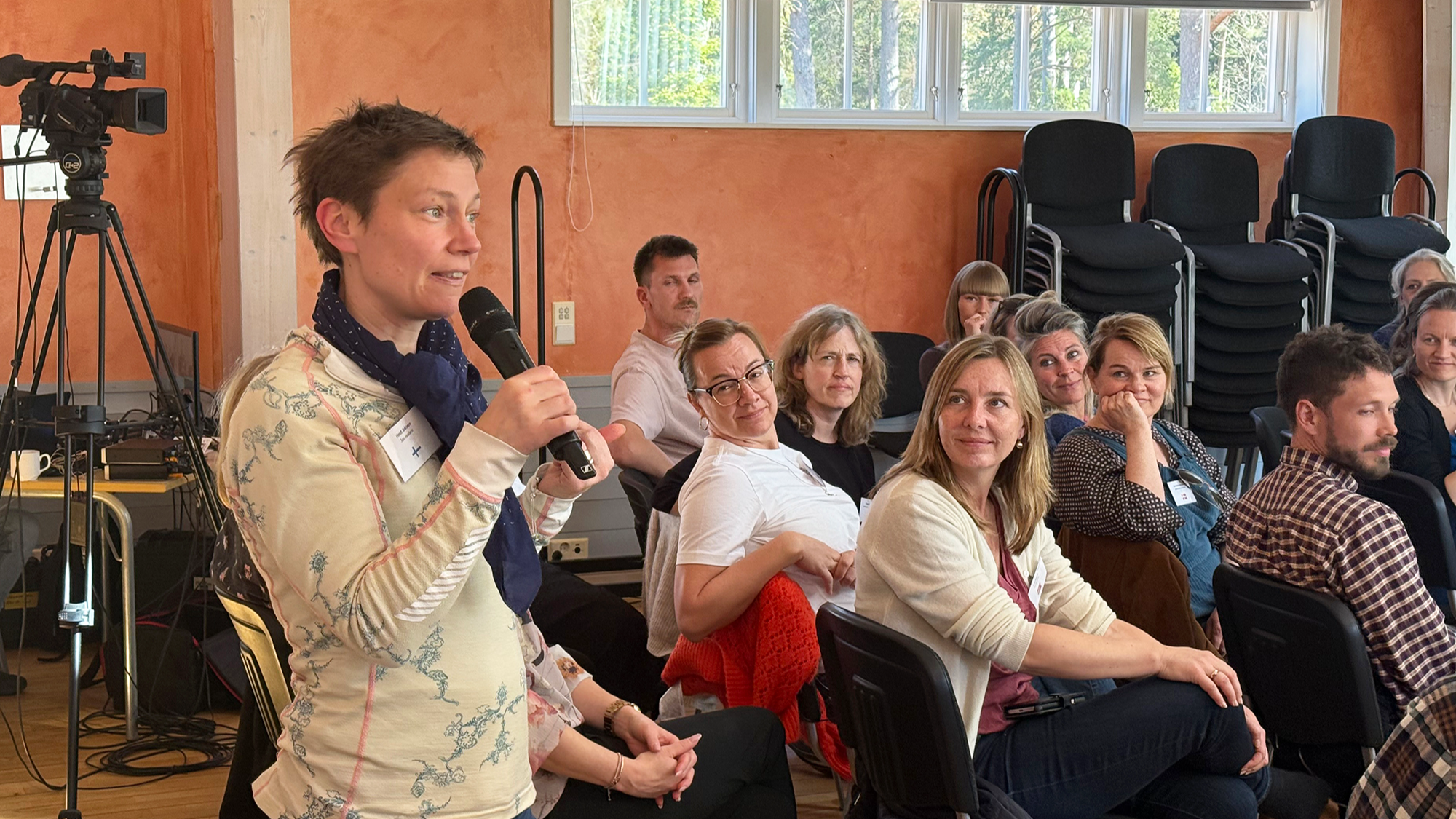 The picture is showing a room with a woman talking on a microphone and the audience is listening. 