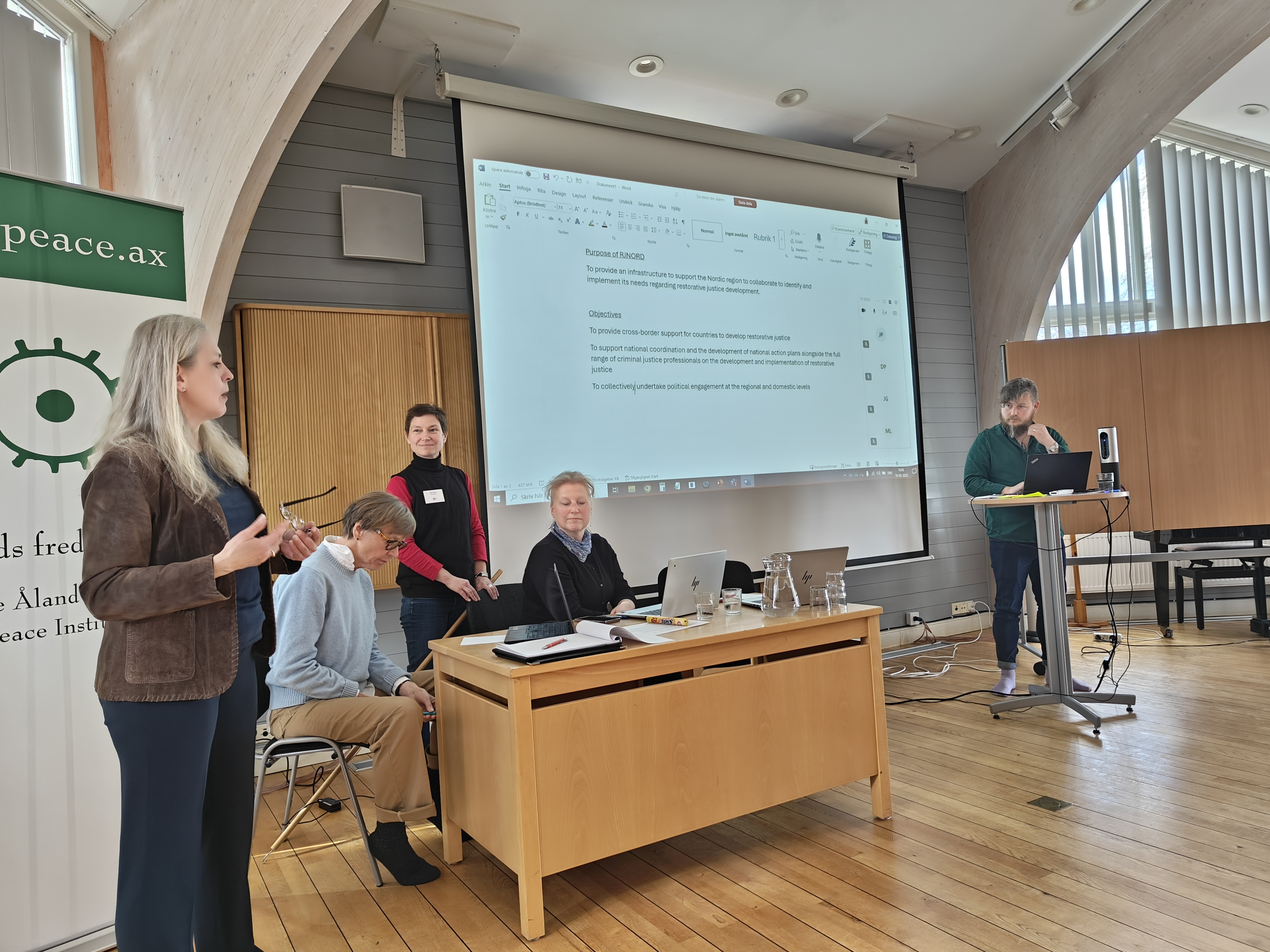 The picture is showing a room with four people standing in front of a screen, one of them writing on a computer. 